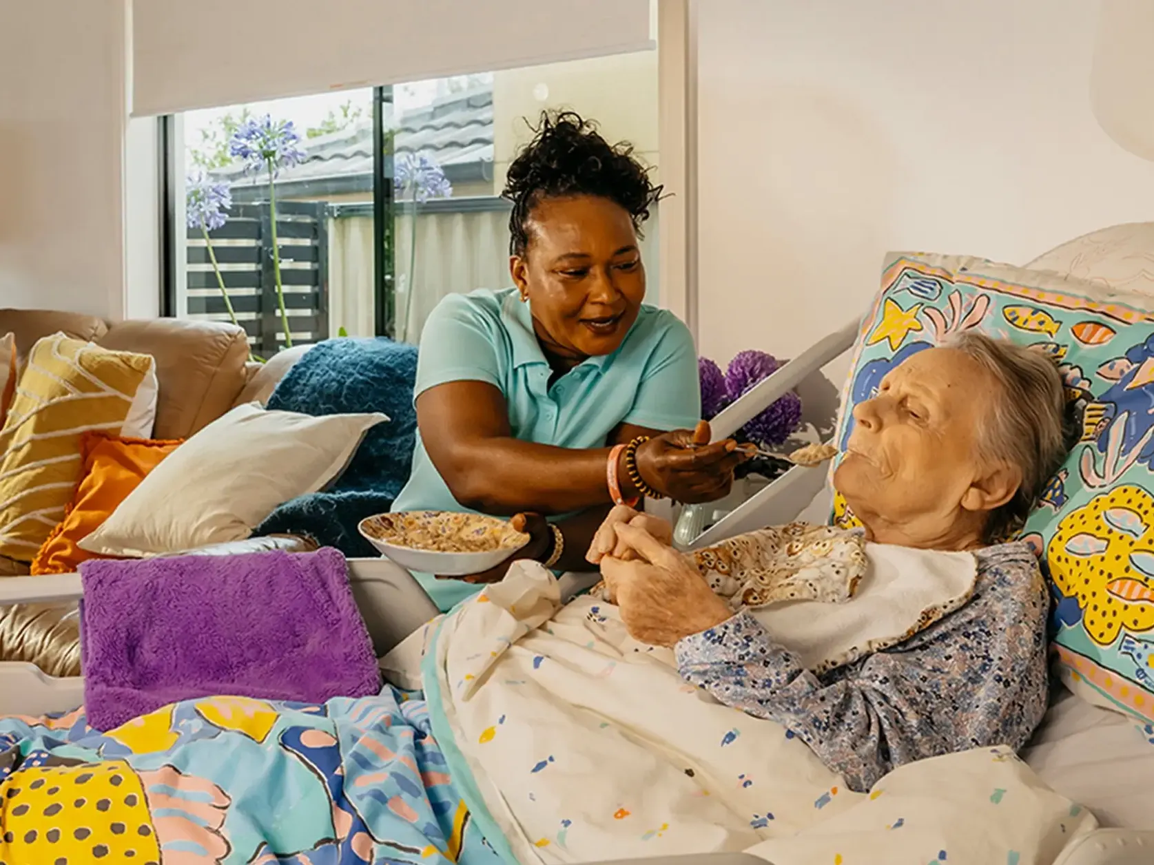 Support worker smiles as they feed an older person living with Dementia laying in bed at home.