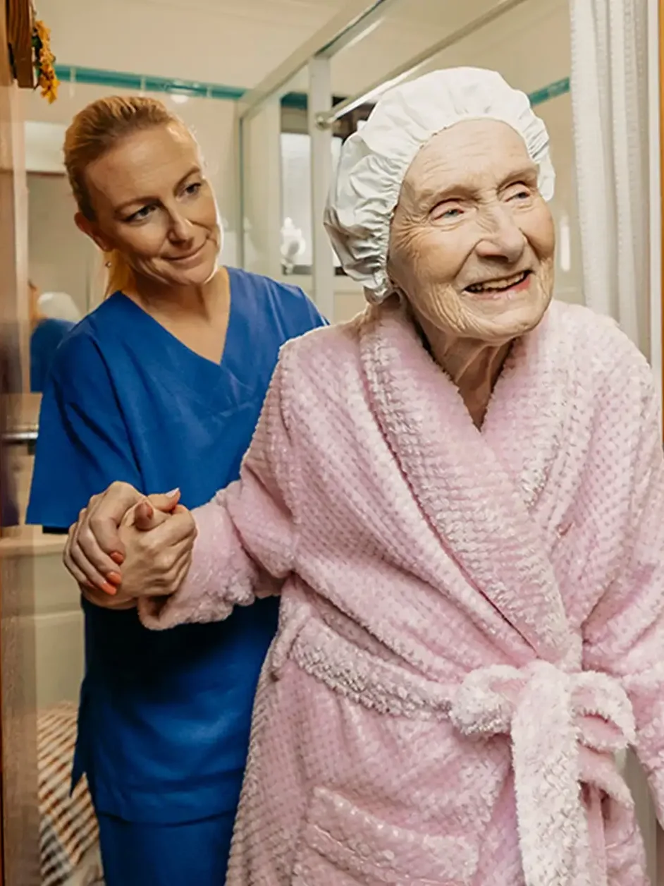 Registered Nurse in scrubs helps older client in pink bathrobe walk out of their bathroom.