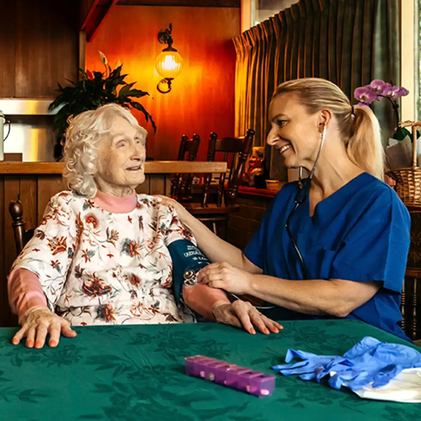 Registered Nurse takes a blood pressure reading for their older client at their dining table. 