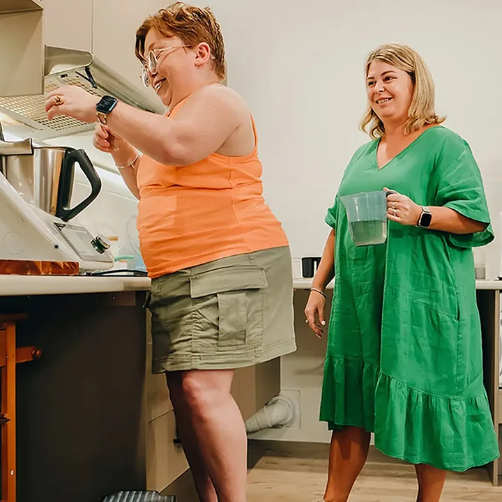 Person with disability stands on step stool at kitchen bench as support worker watches.