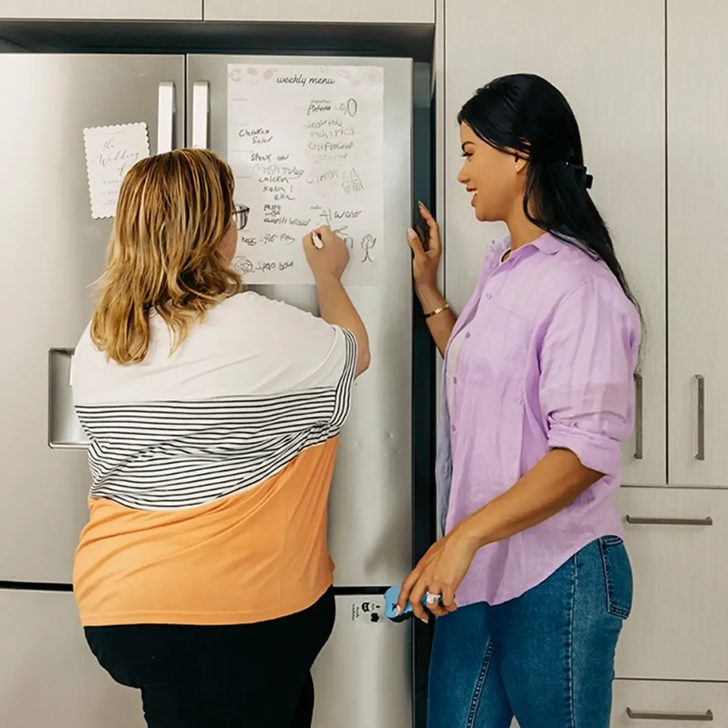 Person with disability in kitchen writes list on fridge while support worker watches.