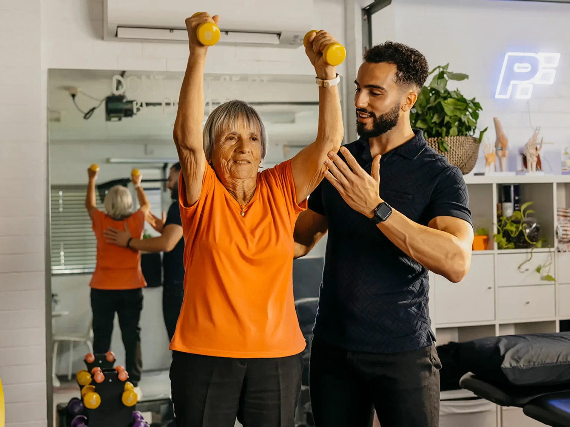 Physiotherapist supports older client’s arm and back as they lift light dumbbells above their head.