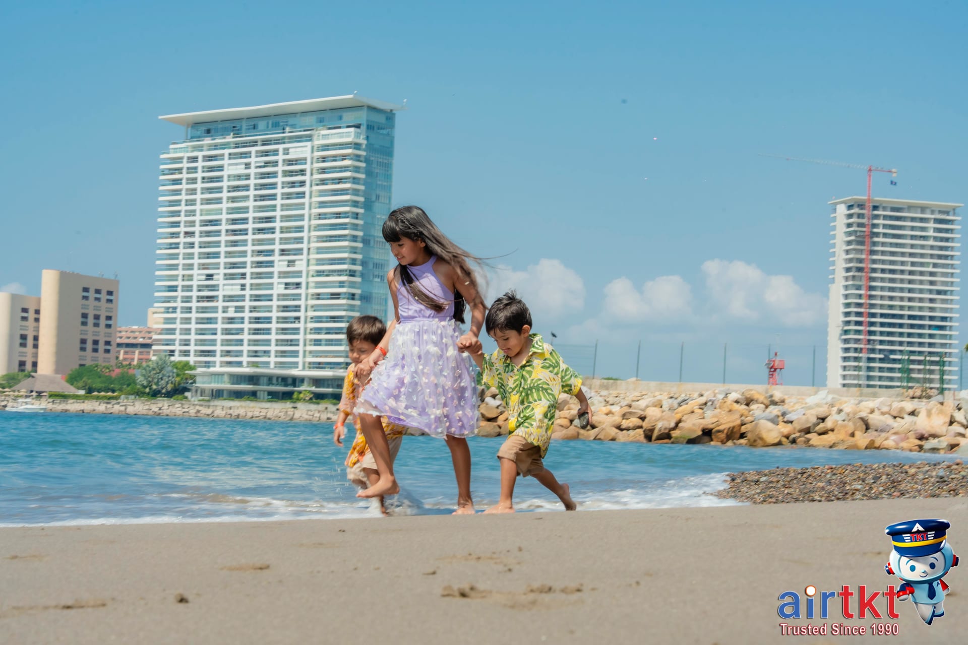 Three children playing on a sunny beach with tall buildings in background in April