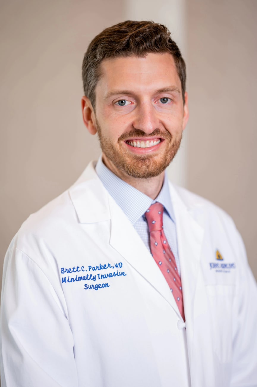 Brett Parker, in a formal portrait, wearing a white lab coat, light blue button down shirt and red tie.