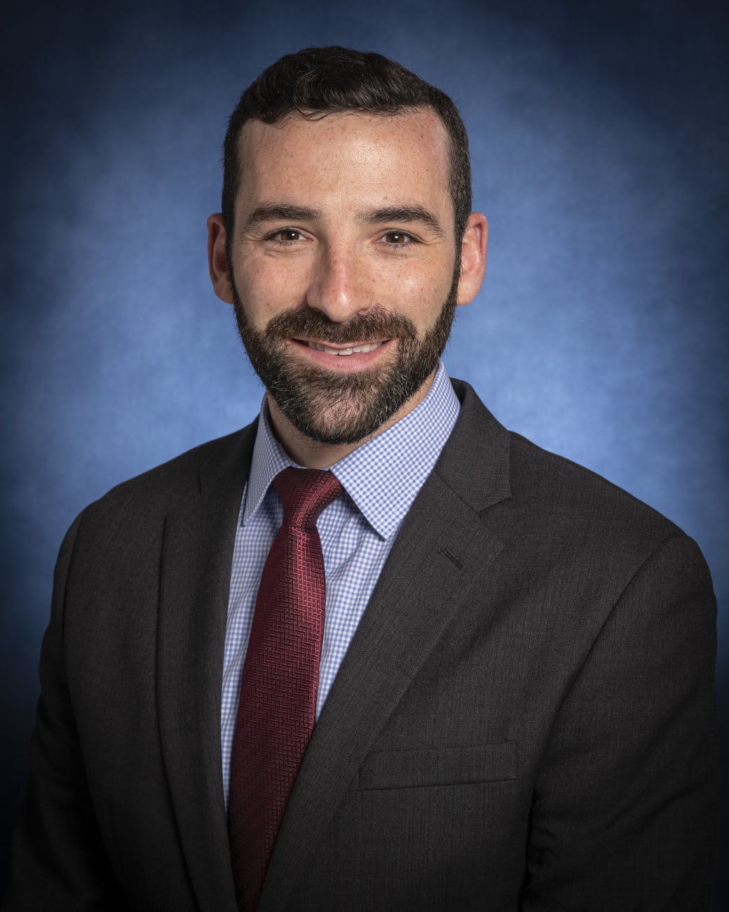 Daniel Lubelski, in a formal portrait, wearing a dark grey suit, blue and white checkered button down shirt and maroon tie. 