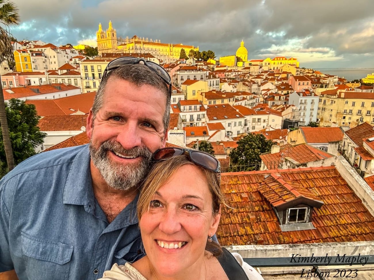 Selfie of a smiling bearded man and a woman, also smiling, with sunglasses on her heard. Red rooftops and a golden sunlit cathedral are in the background behind them.