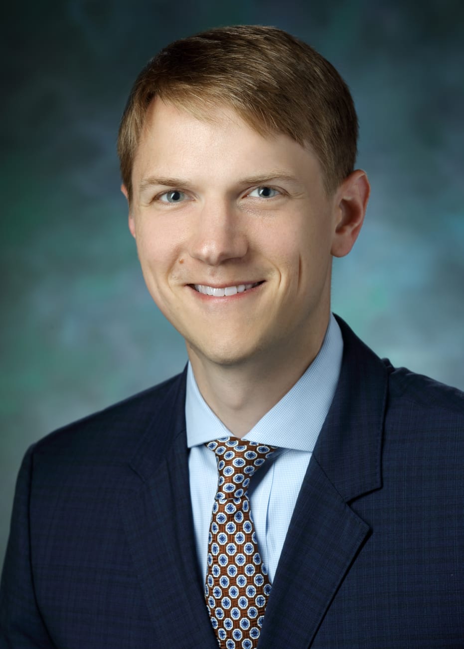 Kelly Mills, in a formal portrait, wearing a dark blue suit, light blue button down shirt and blue tie