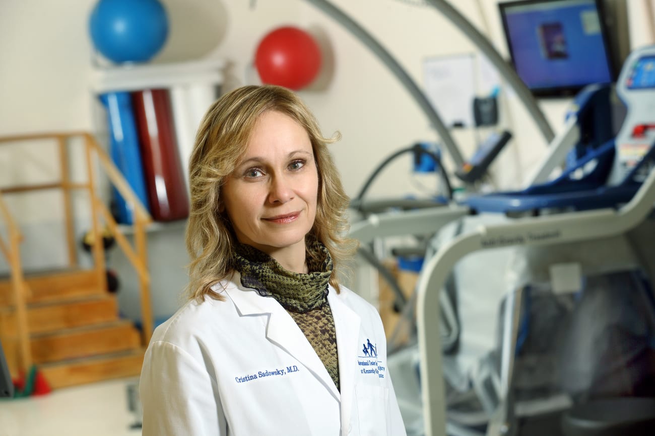 Cristina Sadowsky, wearing a white lab coat, poses in a treatment room with equipment behind her.