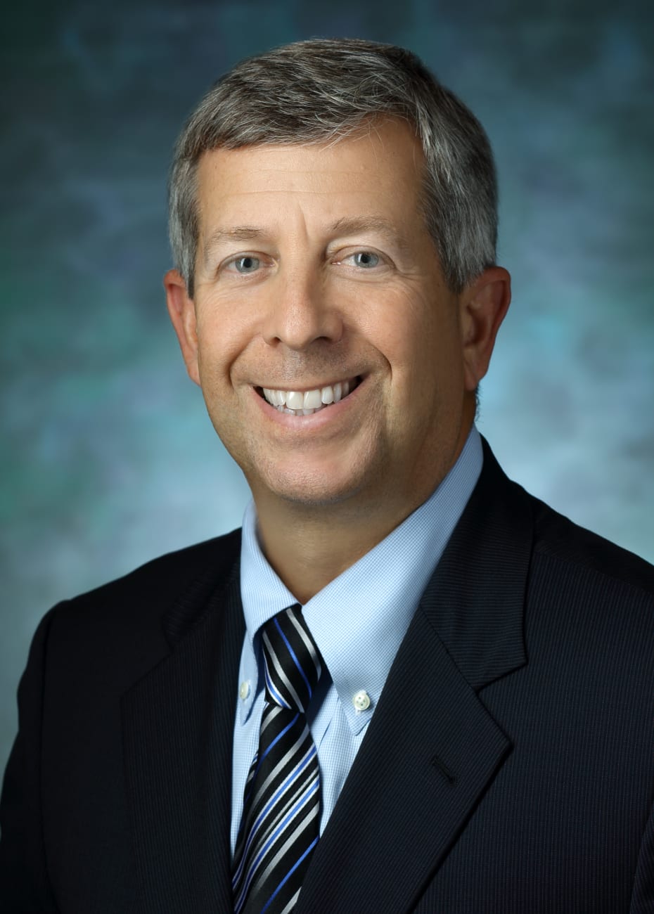 Andrew Satin, in a formal portrait, wearing a blue, silver and black striped tie, light blue button down shirt and dark blue suit jacket