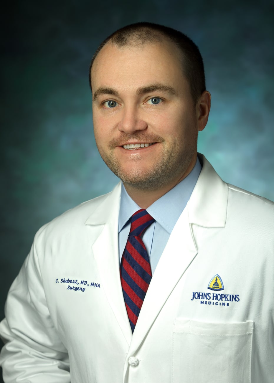 Surgical oncologist Christopher Shubert in a formal portrait, wearing a white lab coat, light blue button down and red and blue striped tie.