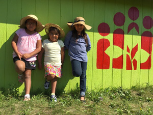 Three indigenous Canadian girls standing by a green wall with a CFTC logo painted on it