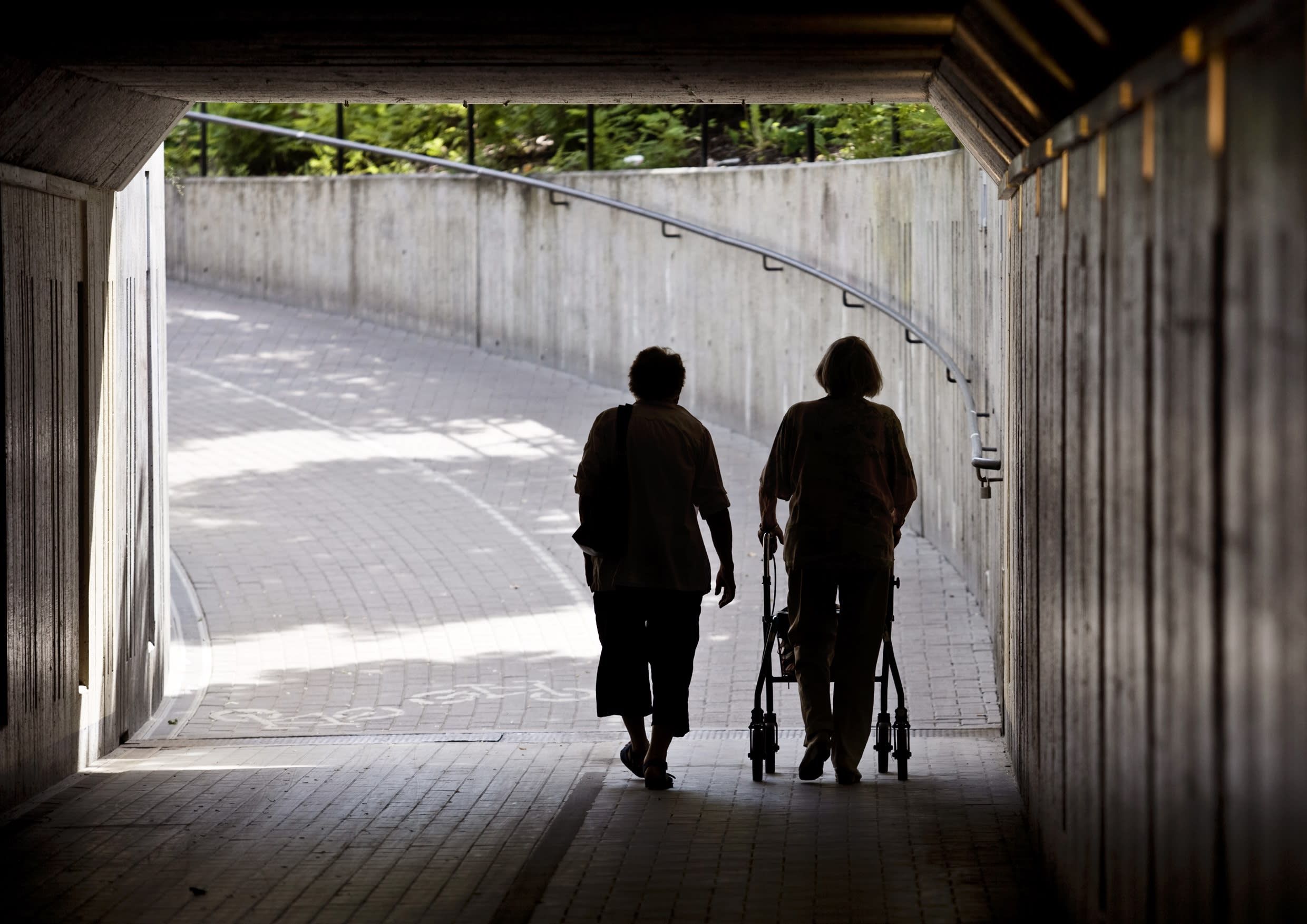 silhuetten av två äldre kvinnor med rollator som går mot ljuset genom gångtunnel i cement