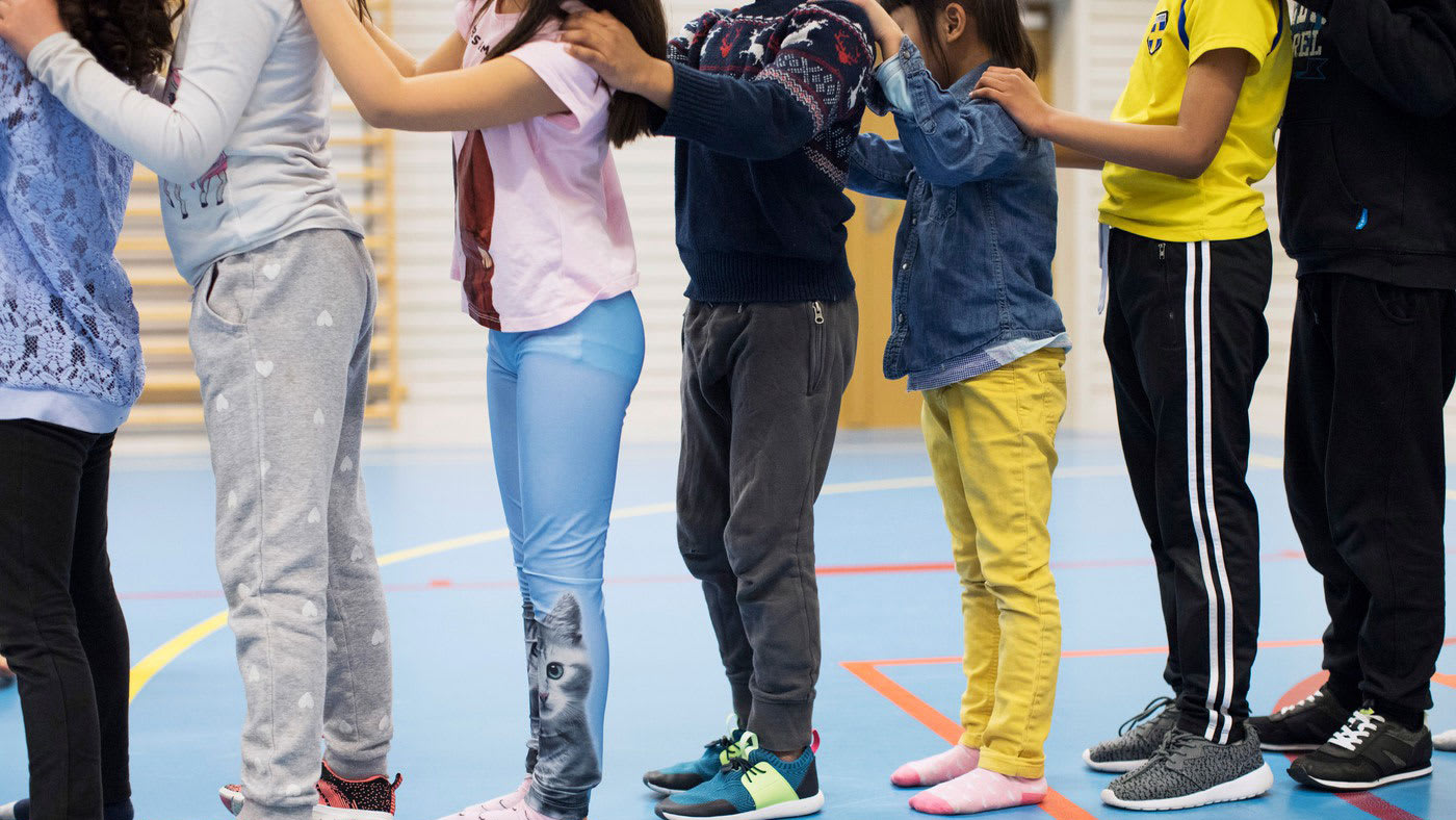 Barn i lågstadieåldern står på led i en gymnastiksal och håller sina händer på varandra axlar i 