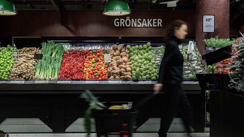 Person med varukorg framför grönsaksdisk i mataffär