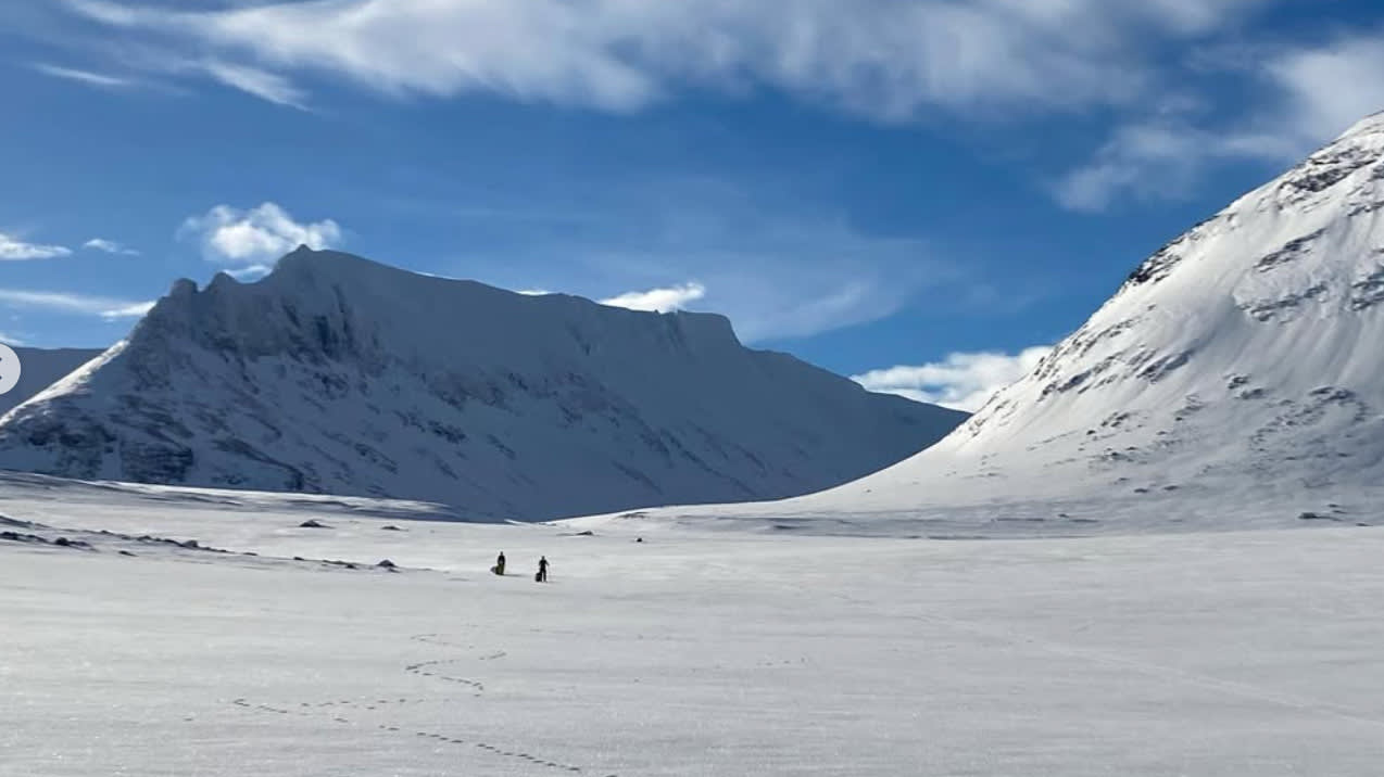 Vitt dramatiskt fjällandskap, solsken och blå himmel, långt bort i bilden skymtas två skidåkare. 