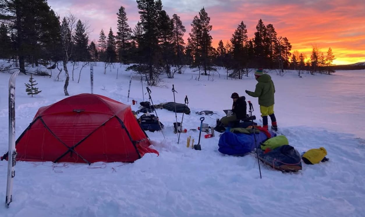 Tält uppspända i snö. Rosaröd himmel, skog och fjäll i bakgunden
