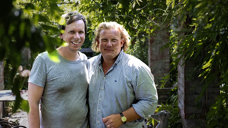 Två medelålders män står på en grönskande innergård