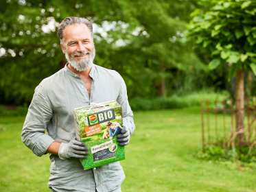 Mann steht auf Rasen und hält eine Schachtel COMPO BIO Rasendünger in der Hand