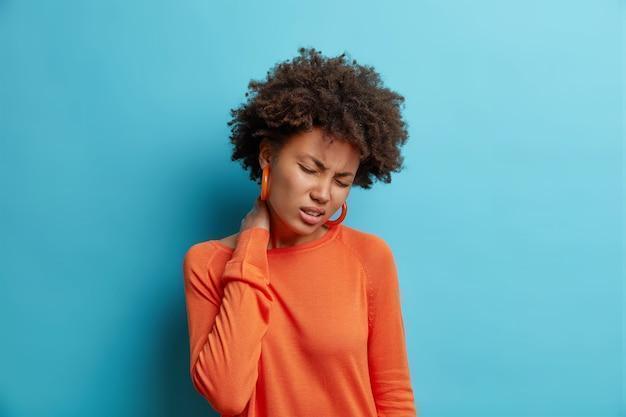 unhappy young woman suffers from pain neck feels tired massages neck feels discomfort closes eyes wears casual orange jumper isolated over blue wall