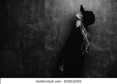 Beautiful young mysterious blond girl in black hat and black jacket on gray background. Eyes are covered with a hat. Manicure - long red nails, nail polish. Fashion, beauty.