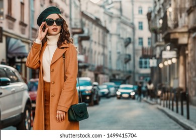 Outdoor autumn portrait of young elegant fashionable woman wearing trendy sunglasses, camel color coat, turtleneck, with textured leather shoulder bag, walking in street of European city. Copy space