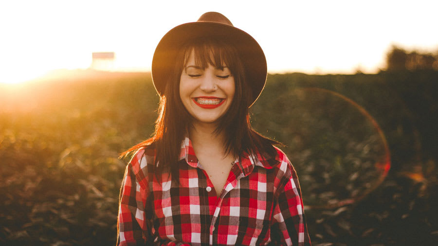 Woman with hat and plaid shirt and red lipstick smiling outside with sunset behind her