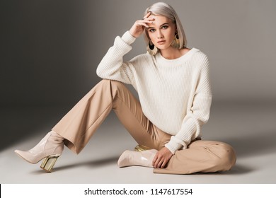 attractive fashionable woman posing in white trendy sweater, beige pants and autumn heels, on grey