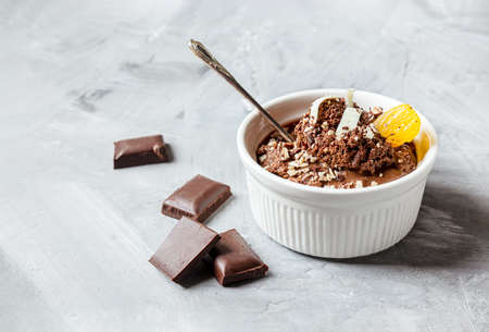 Chocolate mousse garnished with a tangerine wedge and white chocolate shavings in white bowl and pieces of chocolate on a gray table