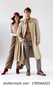 full length view of stylish woman in hat touching hand of trendy man in trench coat on grey