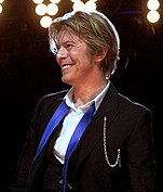 A man smiling; he has brown hair and wears a suit jacket and vest, and a white shirt open at the collar. His blue tie is not fastened.
