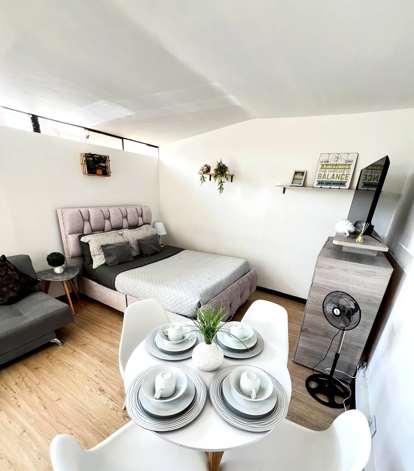 Un espacio de dormitorio elegante con una cama tamaño matrimonial, decorada con cojines y un edredón gris. Hay una mesa pequeña con platos, tazas y un centro de mesa.