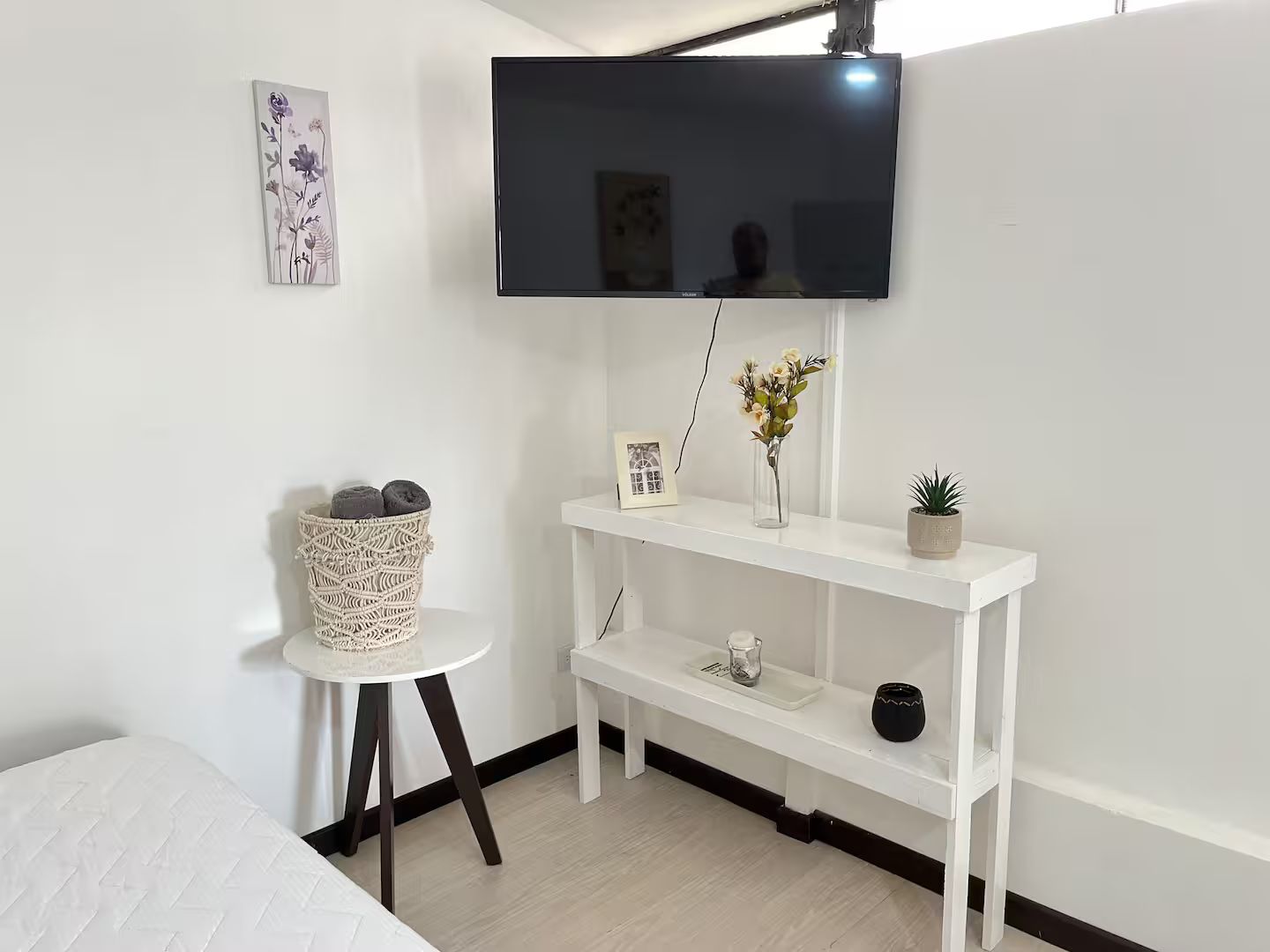 Sala Acogedora: Un televisor montado en la pared sobre una mesa blanca estrecha con decoración mínima. A su lado, una mesa redonda con un cesto decorativo y una decoración de pared con flores.