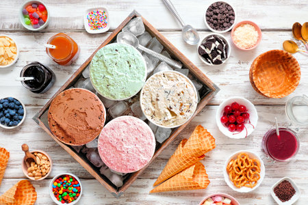 Summer ice cream buffet with a variety of ice cream flavors and sweet toppings. overhead view table scene on a rustic white wood background. Stock Photo