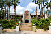The gateway to a facility reading "Jurassic Park" at the top, "Discovery Center" at the bottom, and a rocky panel with a Tyrannosaurus skeleton in-between.