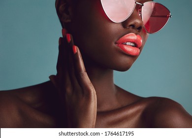 Close-up of a glamorous female model with artistic makeup wearing funky sunglasses. Attractive woman with red lipstick and nailpaint looking away on grey background.