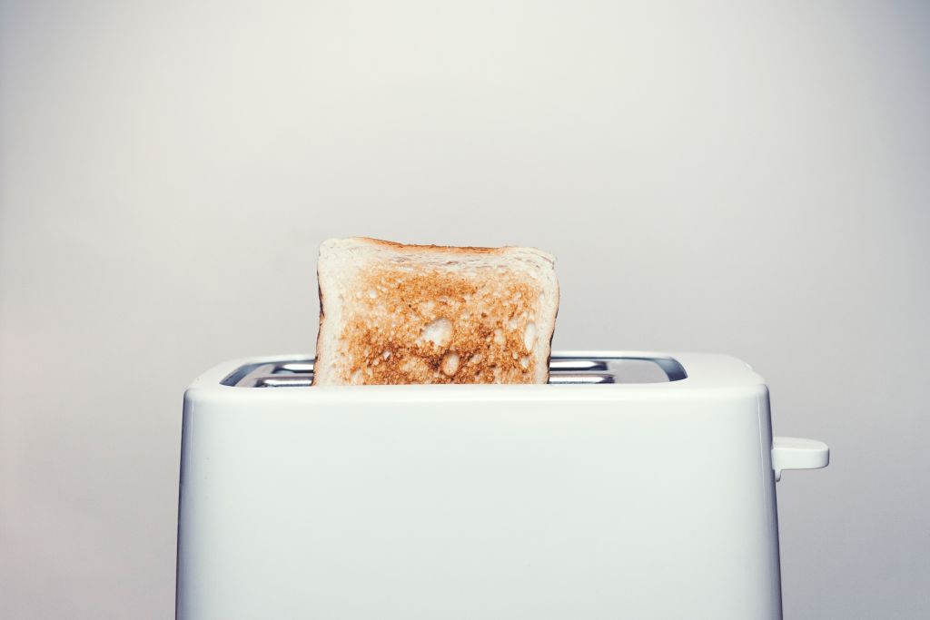 Ein heller Hintergrund mit einem weißen Toaster im Vordergrund. Aus dem Toaster guckt ein fertig getoastetes Toast heraus.