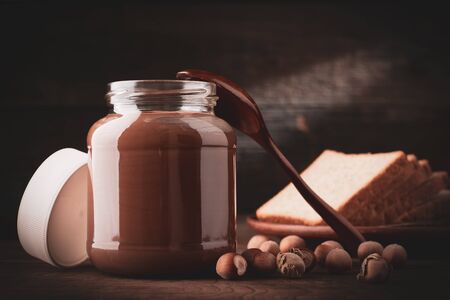 Jar of chocolate-nut pasta with slices of fresh white bread and hazelnuts on wooden background.