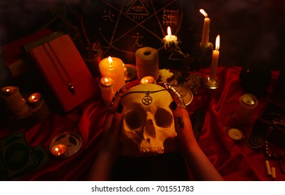 Witchcraft composition with witch's hands holding a human skull, candles, magic books, tarot cards, amulets and pentagram symbol. Halloween and occult concept, black magic ritual.