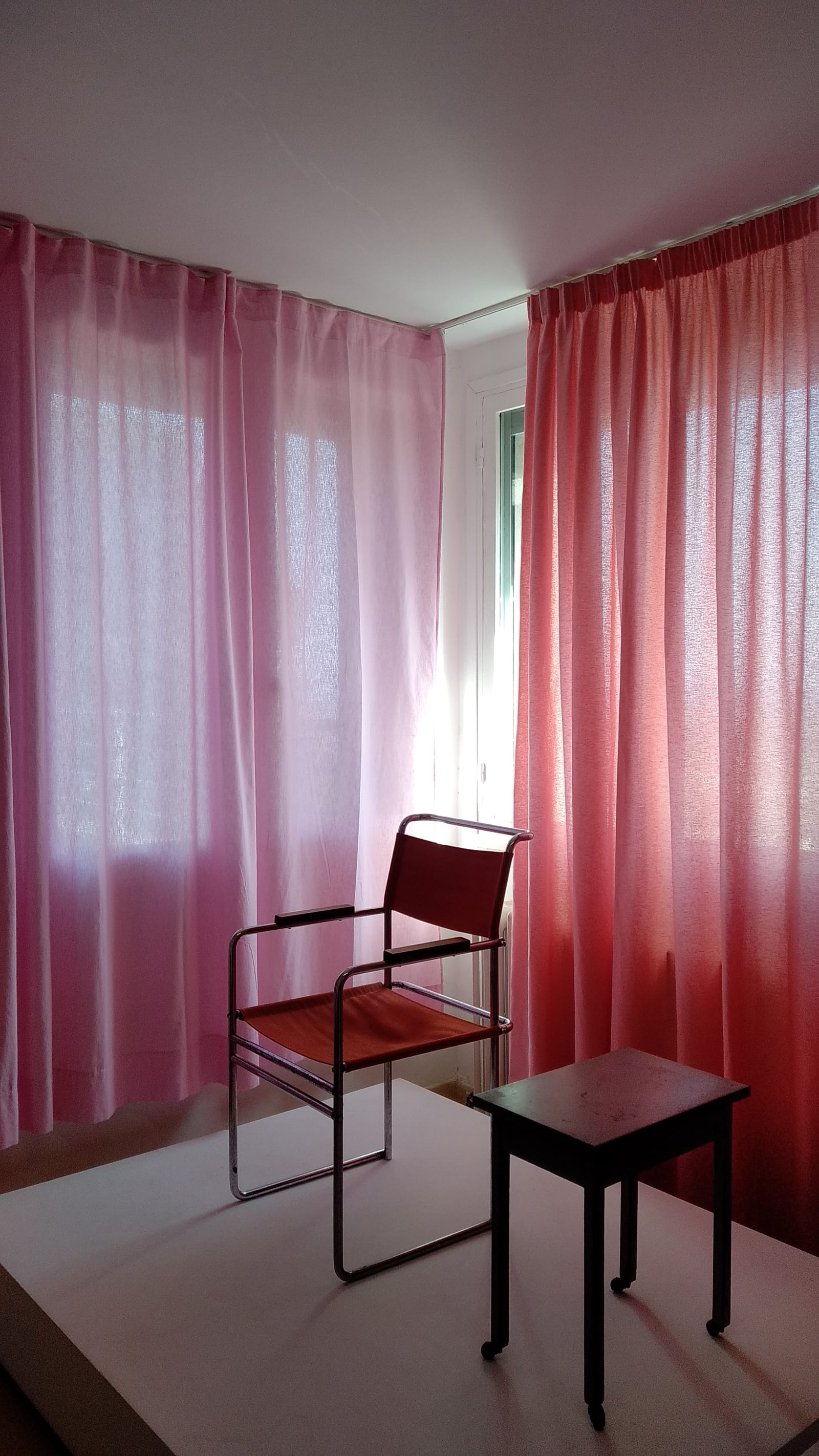 uncertain - In Villa Noailles, a red chair and a brown table are displayed in front of a pink curtain and a coral curtain - chair, table, two (2) curtains - paul lahana