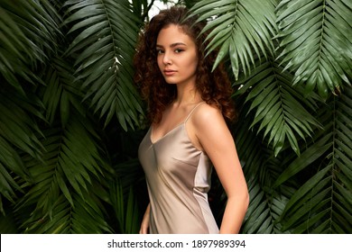 woman in silk dress poses in botanical garden, good-looking lady has natural beauty, stand between green big plants