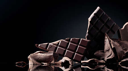 Broken chocolate bar and pieces of dark chocolate on a black reflective background. copy space.