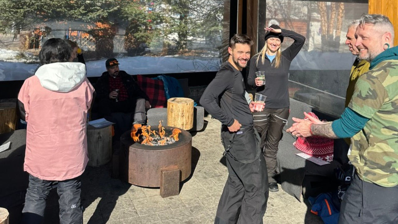 SkiCommerce attendees enjoying an outdoor gathering around a fire pit, with some people chatting, one smiling at the camera with a drink in hand, and another warming their hands by the fire, all against a snowy landscape backdrop.