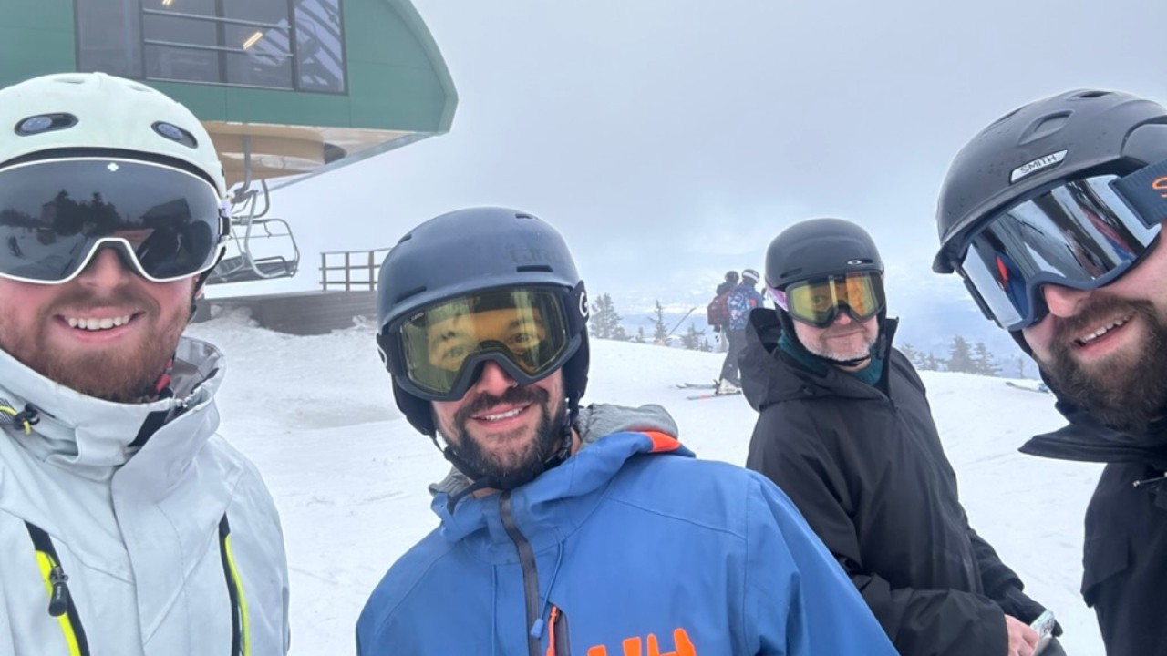 Four smiling SkiCommerce participants pose for a selfie on the ski slopes, donning ski gear and goggles, with a ski lift and snowy mountain in the background, capturing the essence of the event's outdoor activities.