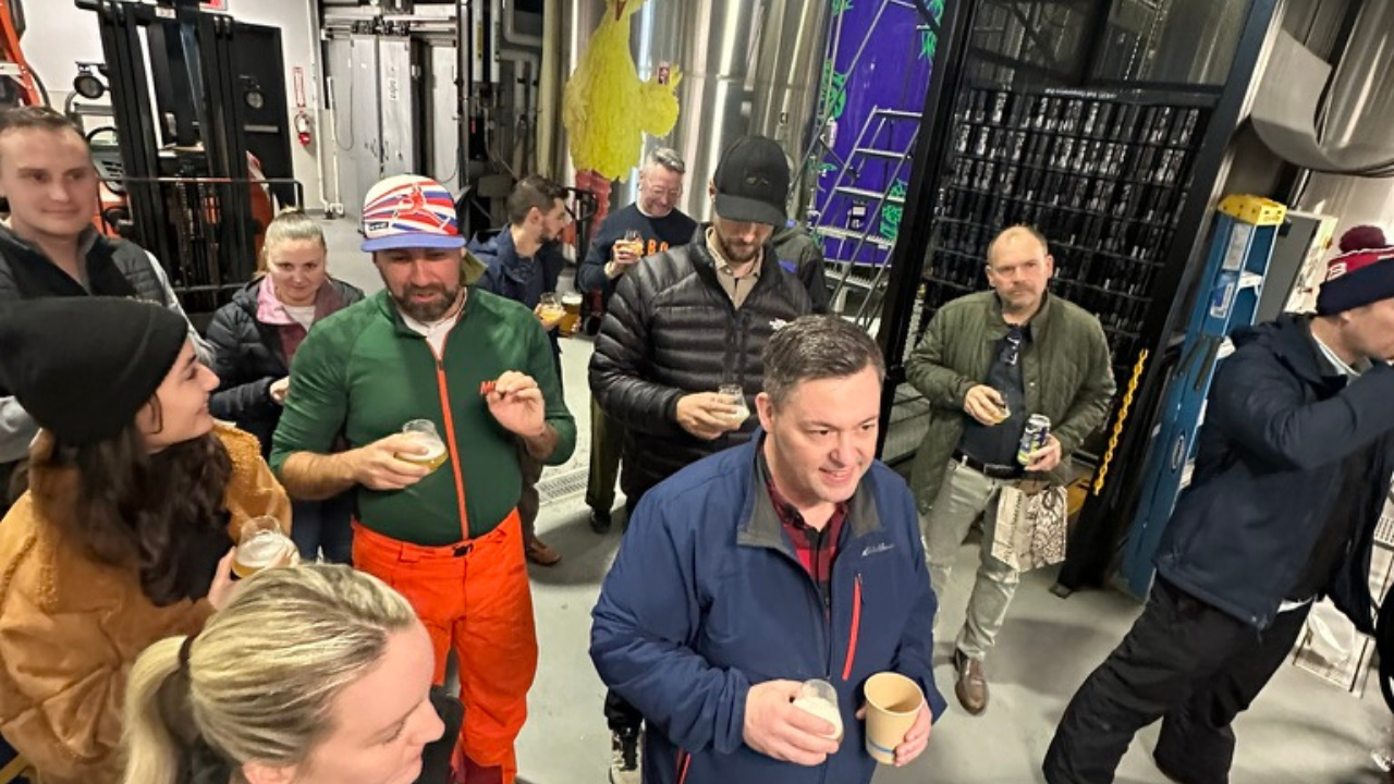 Attendees of SkiCommerce 2024 are engaged in lively conversations while sampling beers during a brewery tour, surrounded by industrial brewing equipment and enjoying the casual, friendly atmosphere.