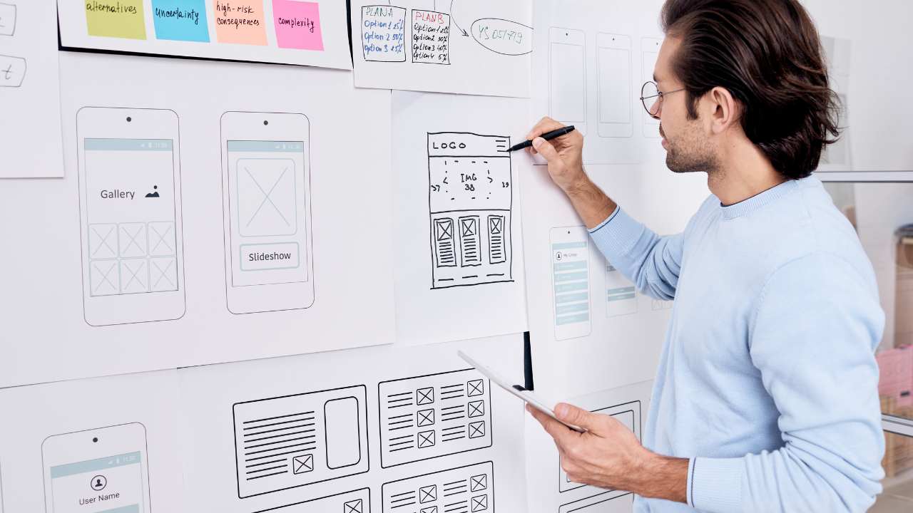 A man with glasses and wearing a light blue sweater is seen from the side, focused on sketching a wireframe for a website or mobile app interface on a whiteboard. The whiteboard is filled with various wireframe designs, sticky notes with annotations, and flowchart elements, indicating a brainstorming or planning session for user interface design.