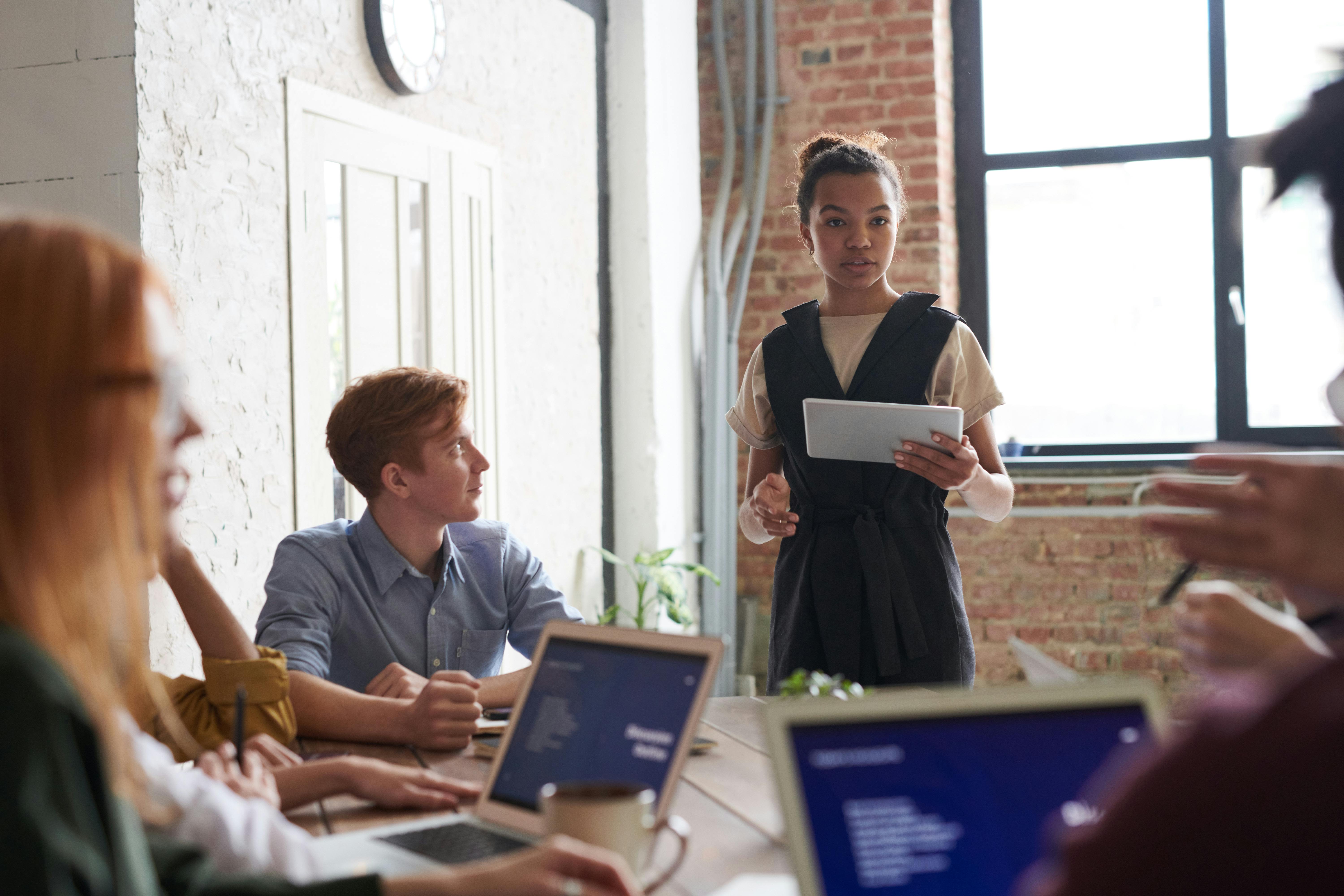 A young professional woman stands confidently at the head of a conference table, holding a digital tablet and addressing an eCommerce marketing team during a meeting. The room is bright with natural light streaming in from large windows, highlighting the casual yet focused atmosphere of a modern workspace with exposed brick walls.