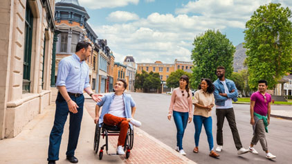 A group of six individuals walks down a sunny, urban street. One person to the left converses with a smiling person in a wheelchair. Behind them, four others walk side by side, engaged in a lively discussion. The background features classic building facades under a clear blue sky.