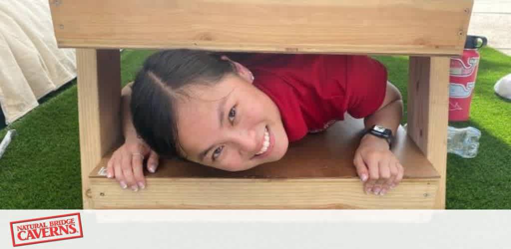 A person smiling while lying inside a small wooden box structure on grass.