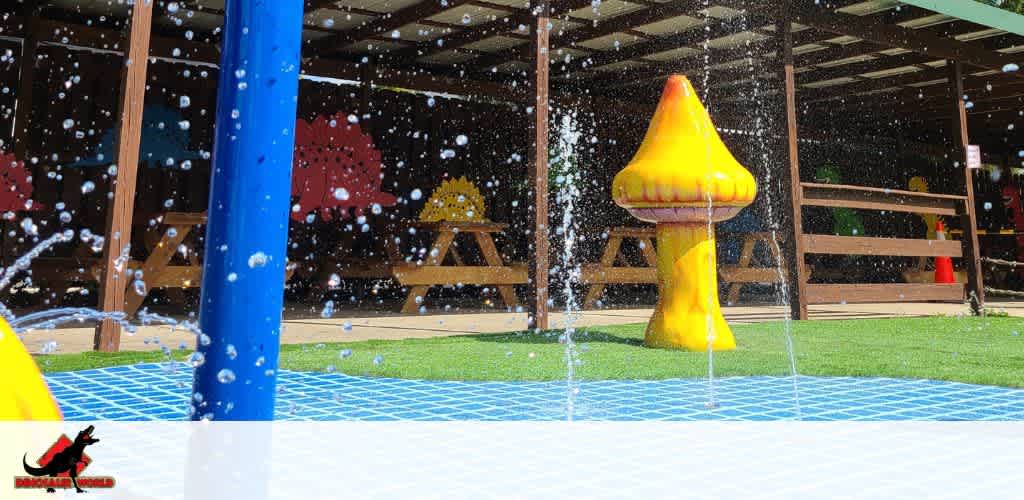 Outdoor splash pad with a yellow mushroom water feature and blue column, picnic tables, and colorful decorations.