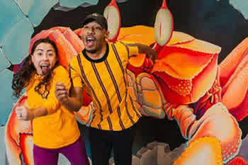 This image depicts two excited individuals inside a room with a whimsical atmosphere. On the left, there is a person with shoulder-length curly hair wearing a yellow top, their mouth wide open in a surprised expression, and their hands up near their cheeks. To the right, another individual is shown wearing a black and yellow striped shirt, also with their mouth open in astonishment, and their eyes wide as they lean towards the left of the frame. Behind them is a large, colorful artwork of a cartoonish red crab with extended claws that appears as though it's chasing after them, adding a playful and dynamic feel to the scene. The backdrop consists of shades of blue that resemble underwater scenery, contributing to the fantasy-like experience. Remember, at FunEx.com, you can always find the lowest prices on tickets, ensuring your adventures are not only memorable but also come with great savings!